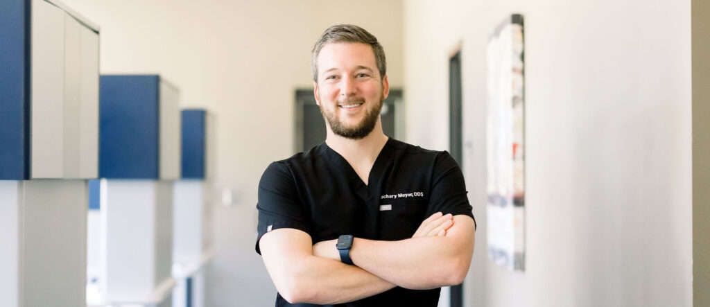 Person wearing a black scrub top with “Zachary Meyer, DDS” embroidered on it, standing indoors with arms crossed and a smartwatch on the left wrist. Background includes blurred elements of a dental office.
