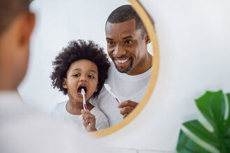 Father and child brushing teeth together in front of the mirror, promoting oral hygiene and healthy dental habits.